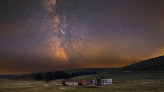 A serene rural landscape at night with an old barn on grassy fields under a starry sky. The Milky Way glows brightly, enhancing the tranquil mood