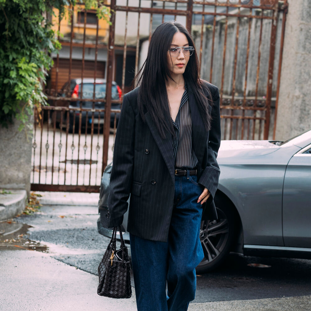 Woman wears jacket, sheer top, jeans, black handbag, and heels. 