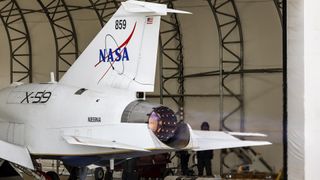 blue and orange flames erupt from the tail end of a white aircraft with a NASA logo on its tail fin while it is parked inside a hangar