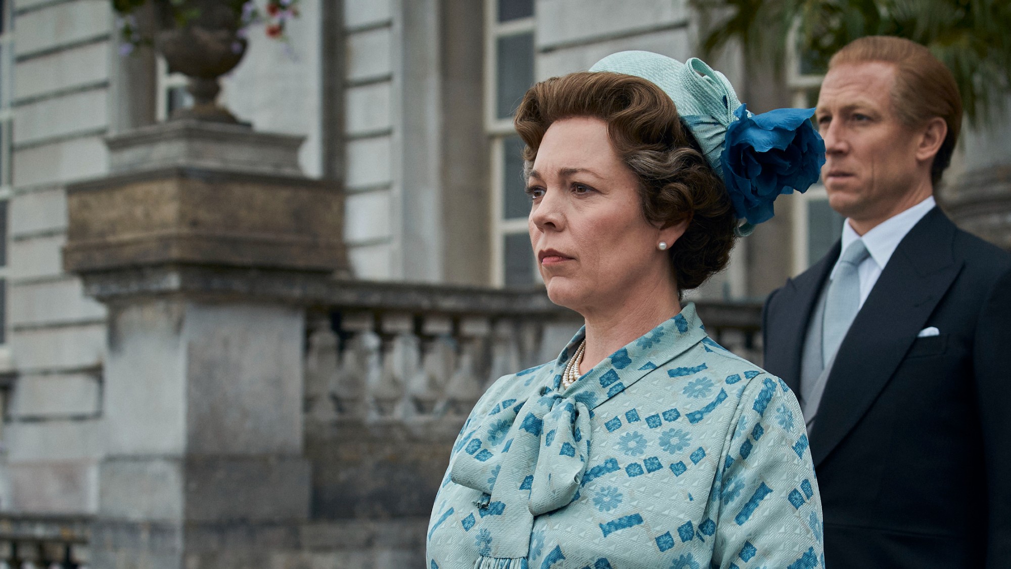straight shot of Olivia Colman as Queen Elizabeth in Season 4 of The Crown. she is dressed in a patterned light blue dress with complimentary hat