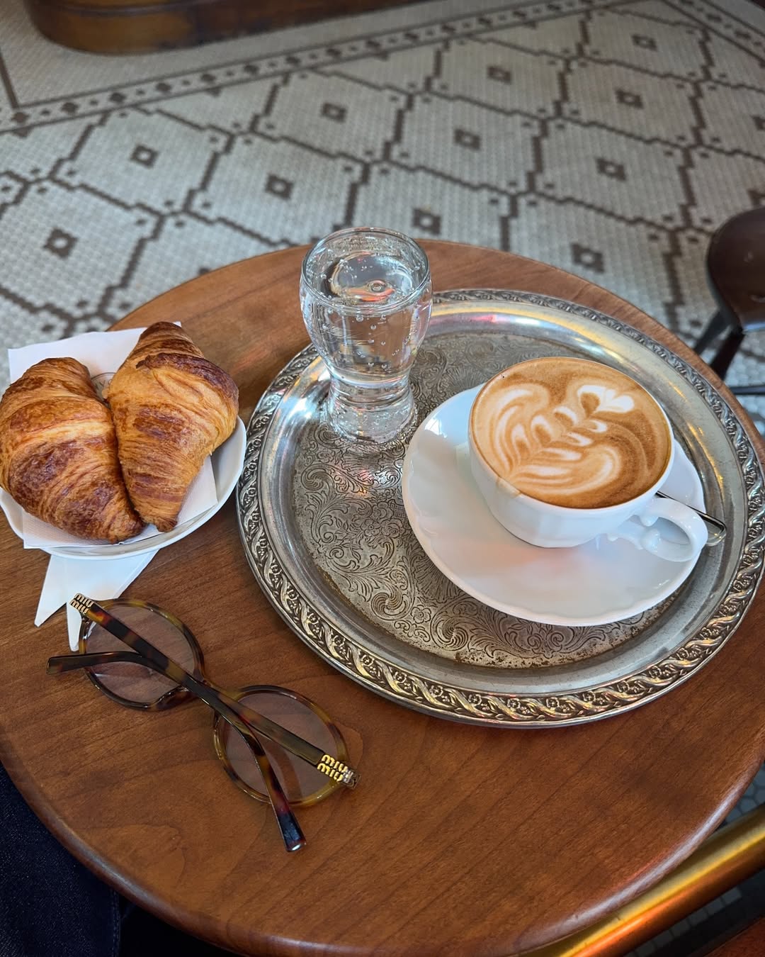 A cafe table with two croissants, water, a latte on a vintage silver platter, and a pair of Miu Miu glasses