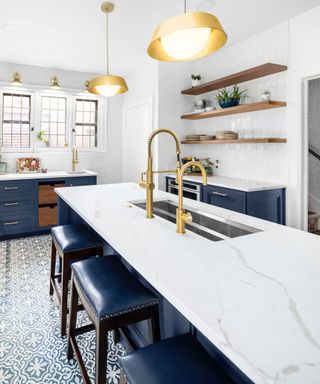 Blue kitchen cabinets with white countertops, white and blue island with integrated sink, brass faucet, gold pendant lights and ornately patterned blue floor tiles