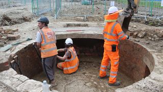 Three archaeologists in orange vests excavating a circular pit.