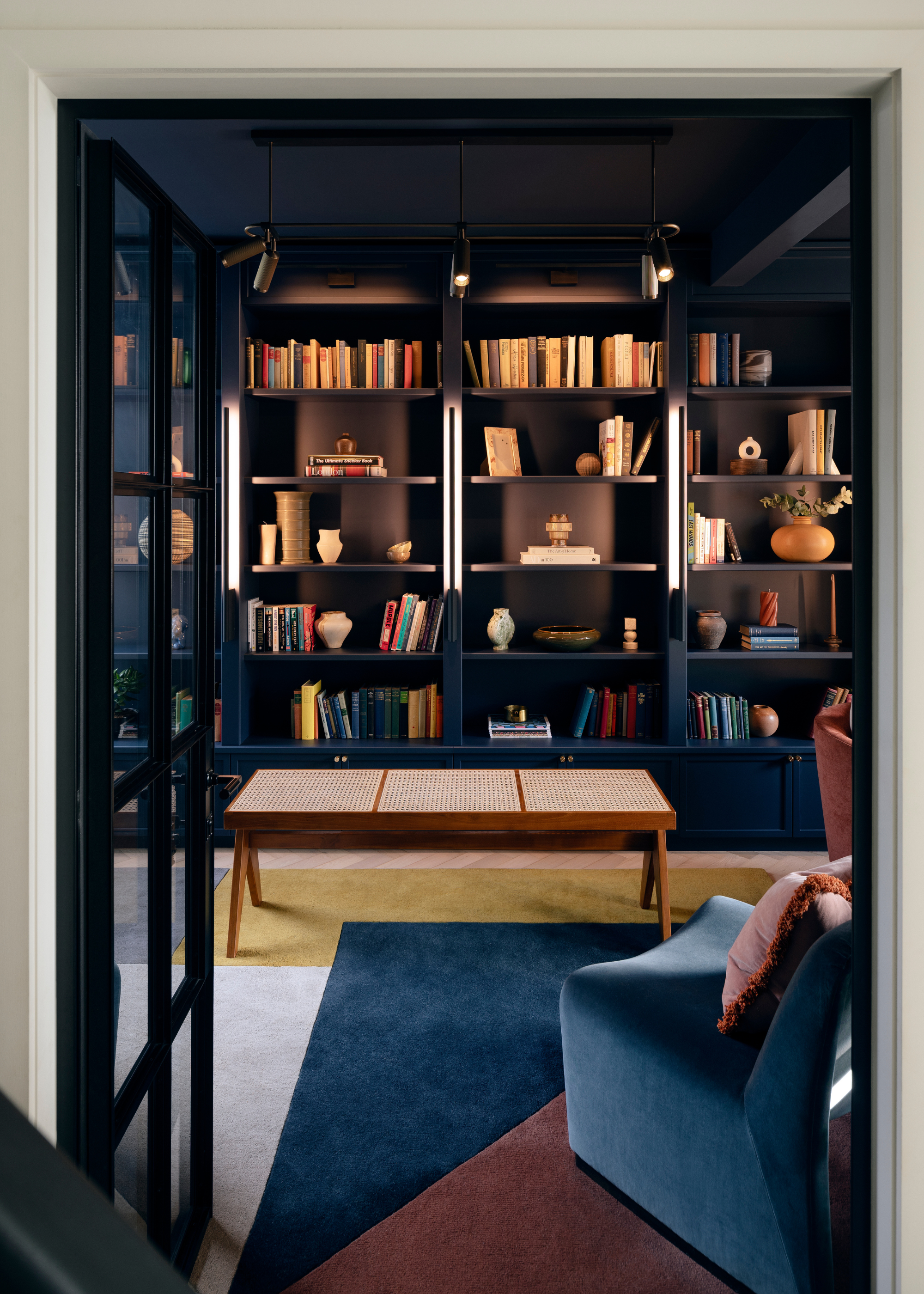 A dark blue reading room with color block carpeting, an illuminated set of storage shelves with books and ceramics, a wood coffee table and blue chairs