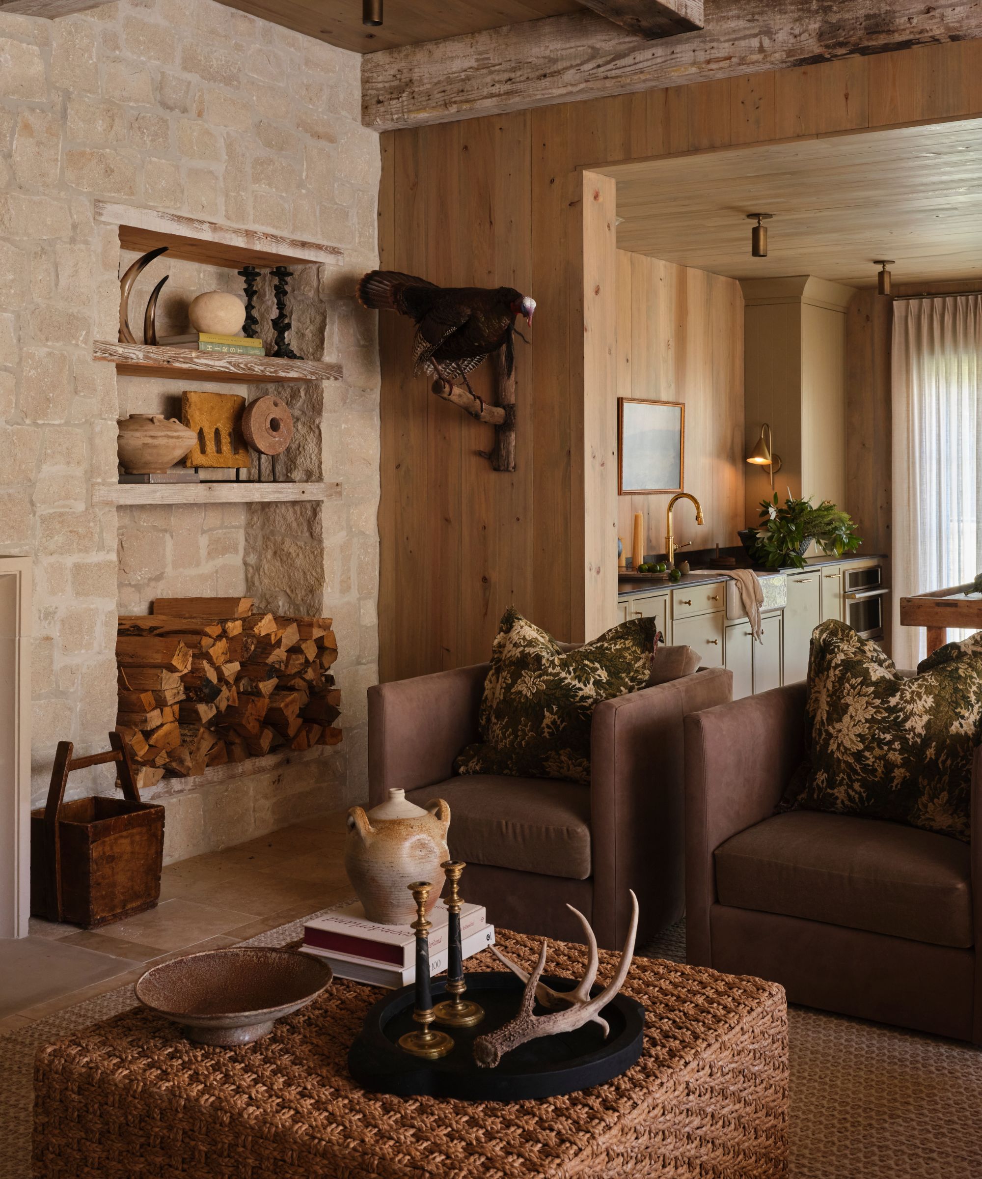 a rustic stone and wood clad living room in a poolhouse with deep purple armchairs, a rattan ottoman and fire wood storage