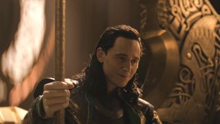 Tom Hiddleston smiles while holding a sceptre as he sits on Asgard's throne in Thor: The Dark World.