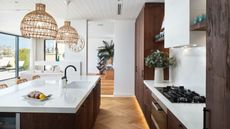Bright, open kitchen area with kitchen island and gas cooker on white countertops. Brown cabinets, hanging wicker lamp shades and a flower vase are also pictured. In the background is an entryway leading to another room with a diagonal bookshelf and large plant.