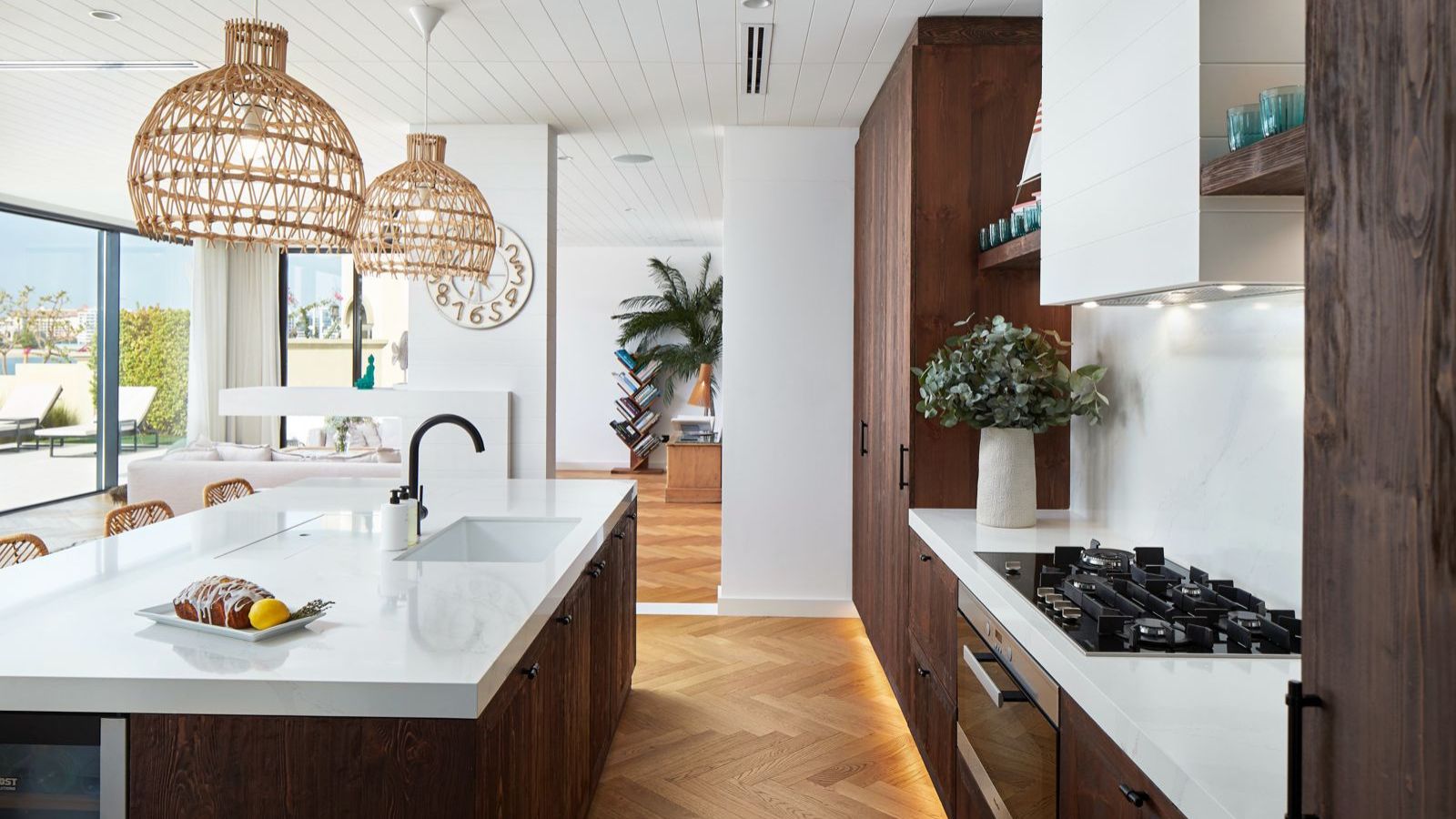 Bright, open kitchen area with kitchen island and gas cooker on white countertops. Brown cabinets, hanging wicker lamp shades and a flower vase are also pictured. In the background is an entryway leading to another room with a diagonal bookshelf and large plant.