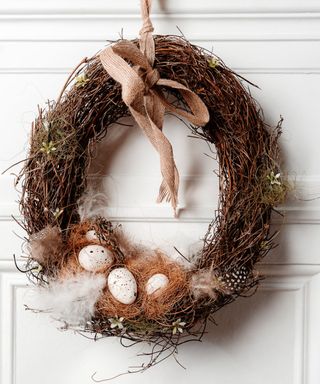 A dark brown twine oval shaped Easter egg with three pastel eggs and feathers at the bottom and a pale pink ribbon tied at the top hanging it up against the white paneled wall