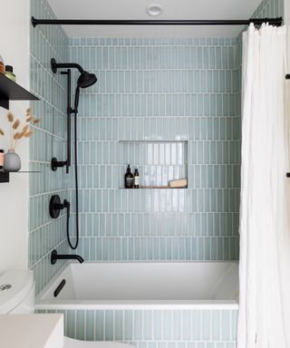 A bathroom with pale blue vertical rectangle tiles around bathtub and shower area, with black faucet and shower hardware