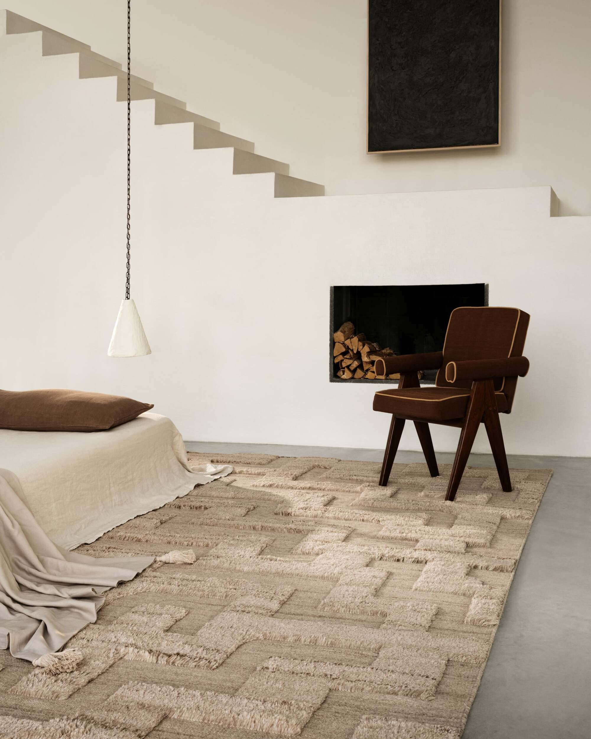 beige tufted rugs with bedcover, low-hanging pendant light, brown armchair in front of a cut-out in the wall with timber, and stairs with an artwork