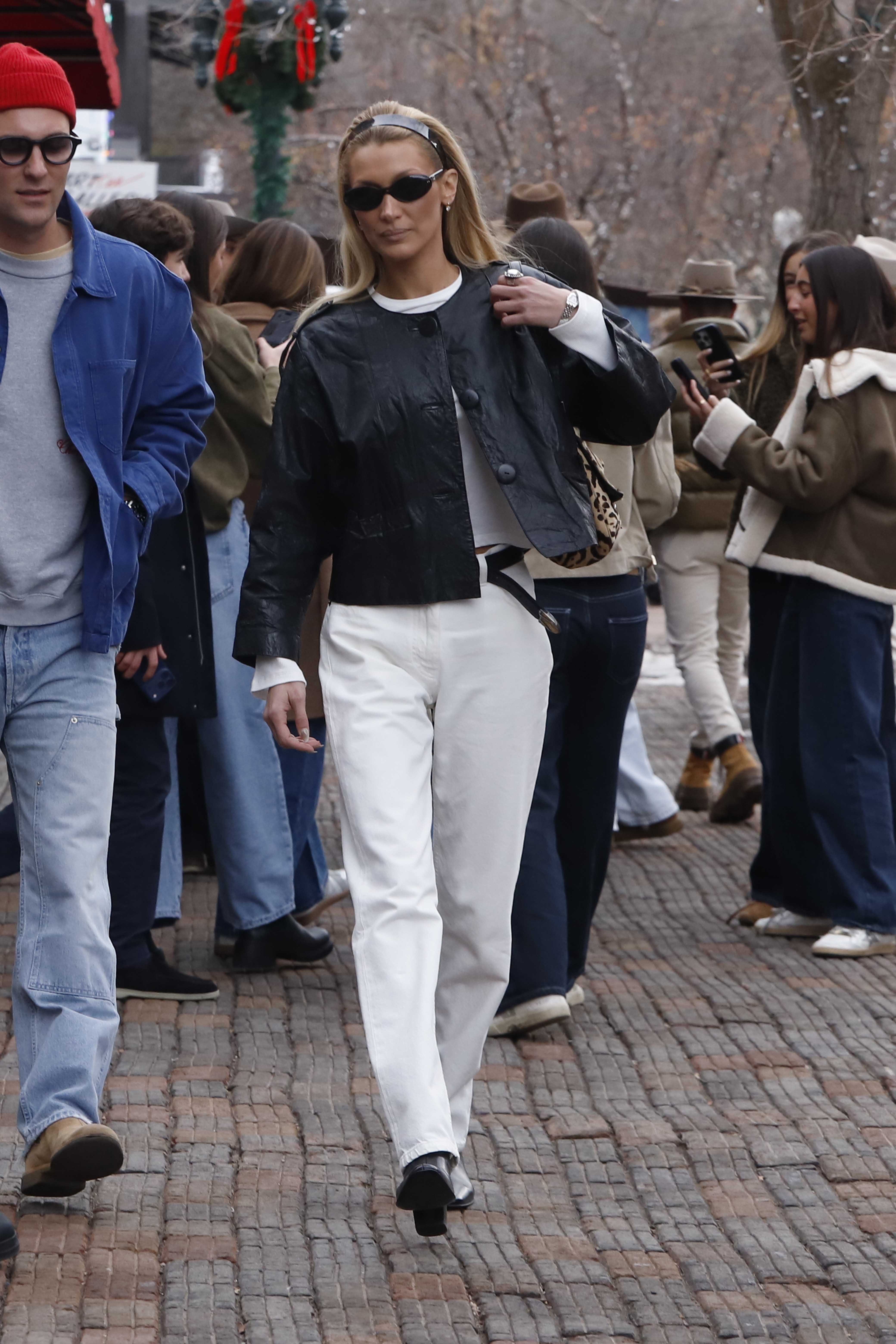 Bella Hadid walks along the pavement outside wearing white jeans with black boots and a black leather jacket.