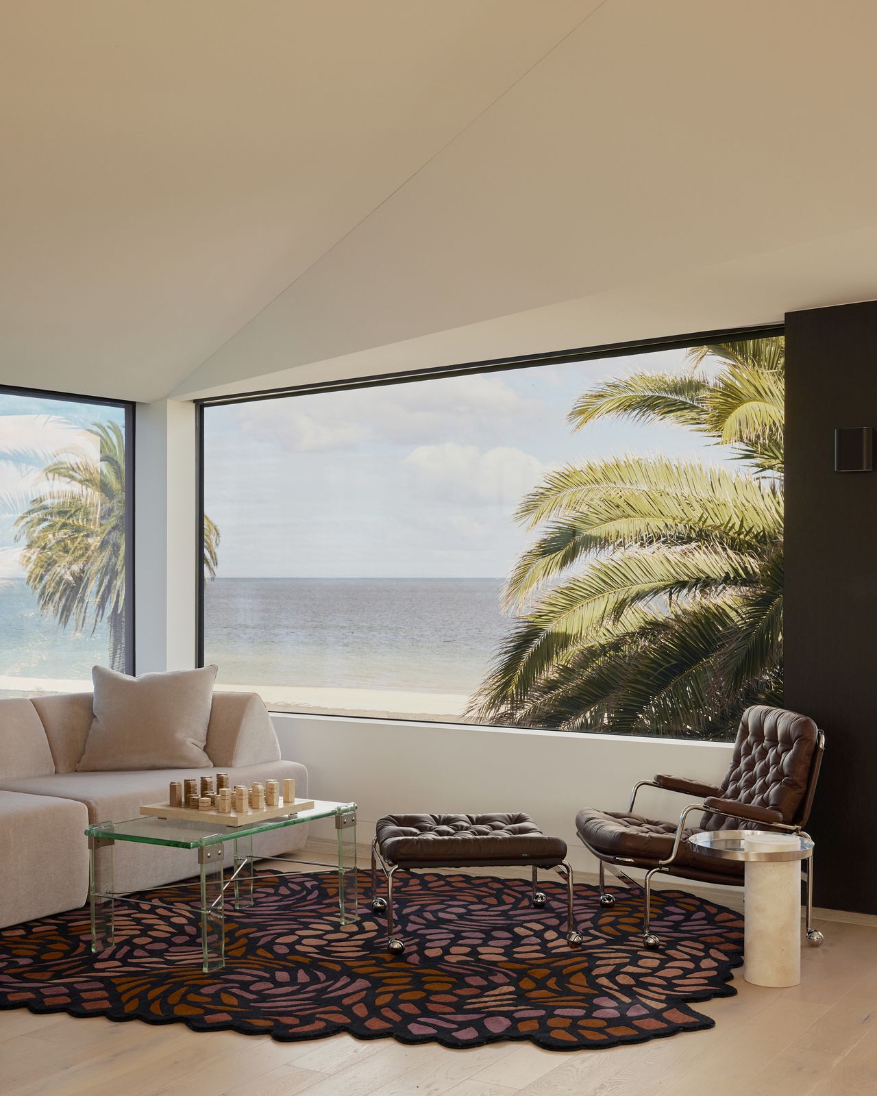 modern coastal living room with big windows with views of palm trees, a leather armchair and matching ottoman, beige sofa, glass coffee table with a chess board, and irregular shaped rug