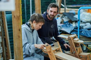 Two people sat at wooden workhorses shape a piece of wood