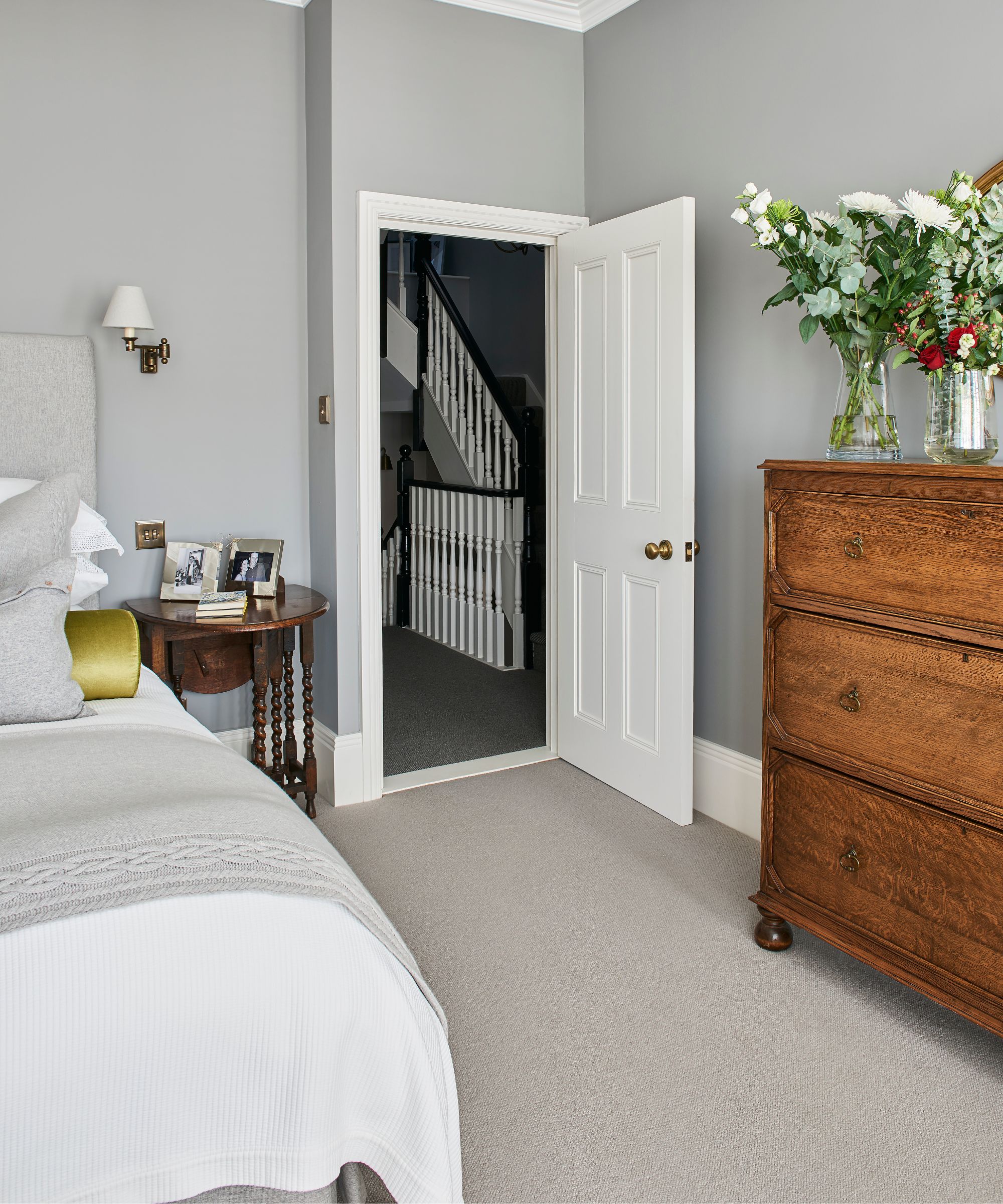 A grey bedroom with a double bed with white bedding seen to the left, a brown bedside table with framed photographs, grey walls and a large brown wooden dresser with glass vases of flowers. The bedroom door is open to show a black and white balustrade behind.