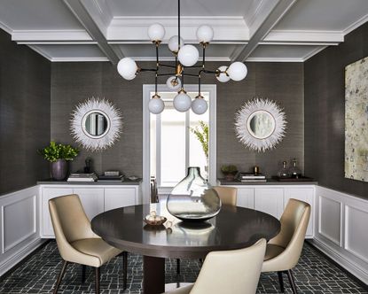 Dining room with white painted white coffered ceiling panels, gray-brown wallpaper, two white rounded mirrors, white wall paneling, black and white, rounded geometric pendant light, round dark wood dining table with four leather dining chairs, square geometric patterned black and white carpet