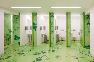 Four bright white WC cubicles in school bathroom. The floor and walls have a flowing dark green leaf print with light green background.