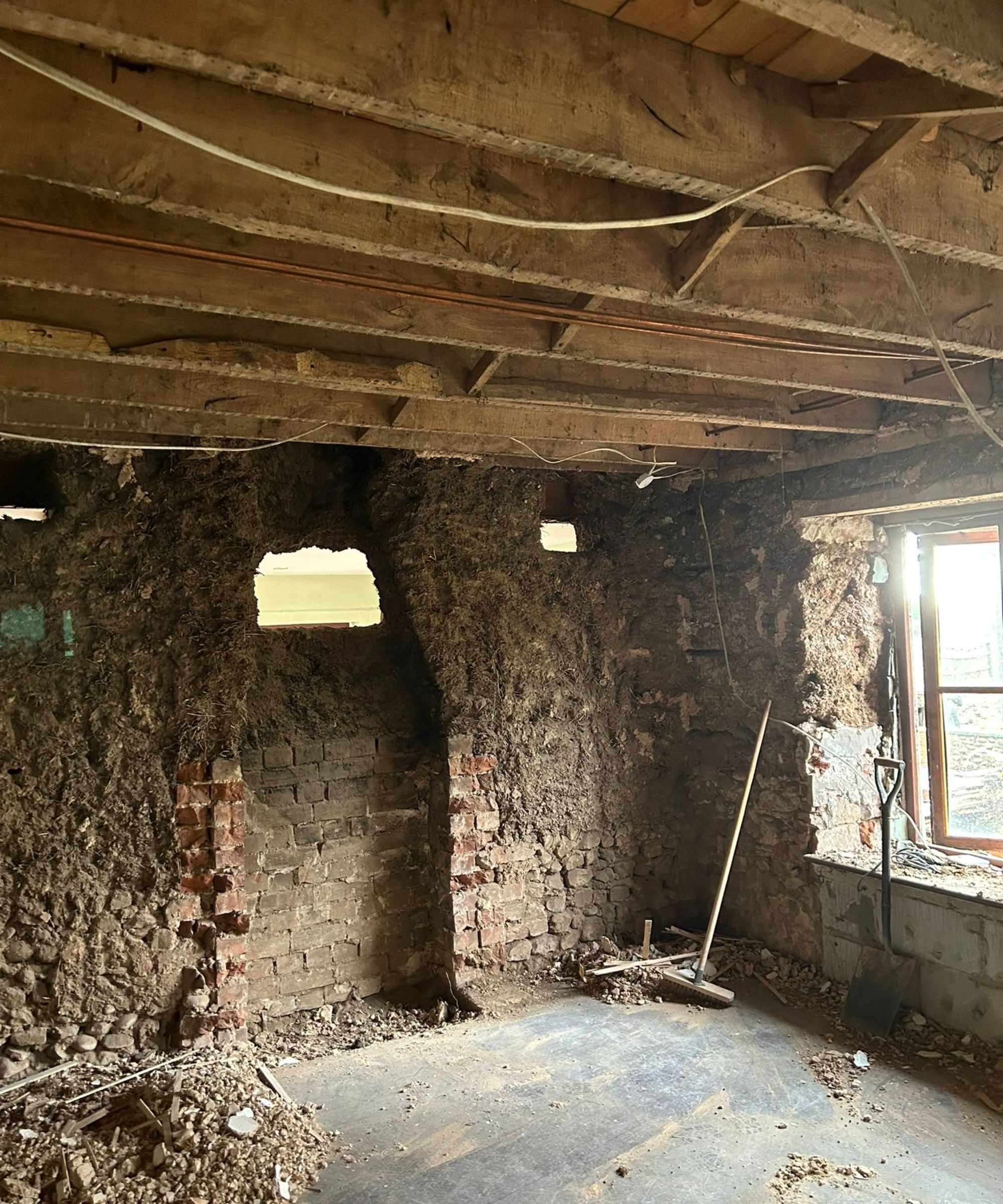 A room mid-renovation with exposed timber ceiling joists, rough cob and brick walls, and rubble scattered on the floor.