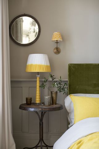 stone bedroom with green upholstered bed, yellow and white bedding, yellow and white table lamp with wall light with yellow lampshade. Vintage side table, vintage mirror, panelling