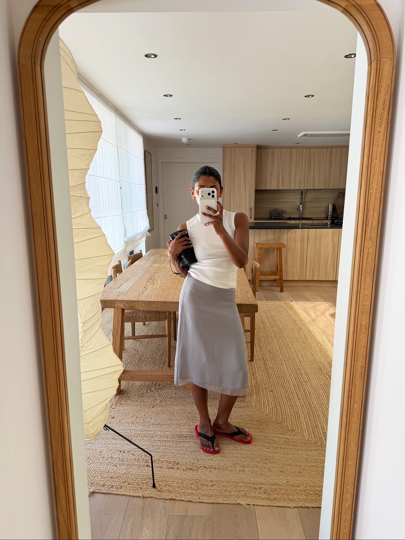 Monique June wears Outfit Ideas 2026. She wears a white tank top, gray midi skirt, and a contrasting red and black pair of The Row Dune sandals. In the recent picture she is holding a leather clutch bag.
