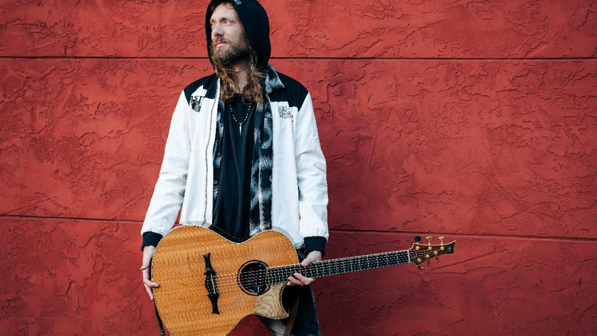 Mike Dawes is photographed wearing a white-and-black hooded top, hood up, against a red background, and holds his custom built cutaway acoustic guitar.
