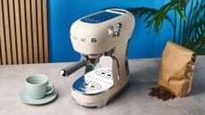 a cream-colored retro espresso machine by italian brand smeg is photographed against a blue background with a bag of coffee beside it