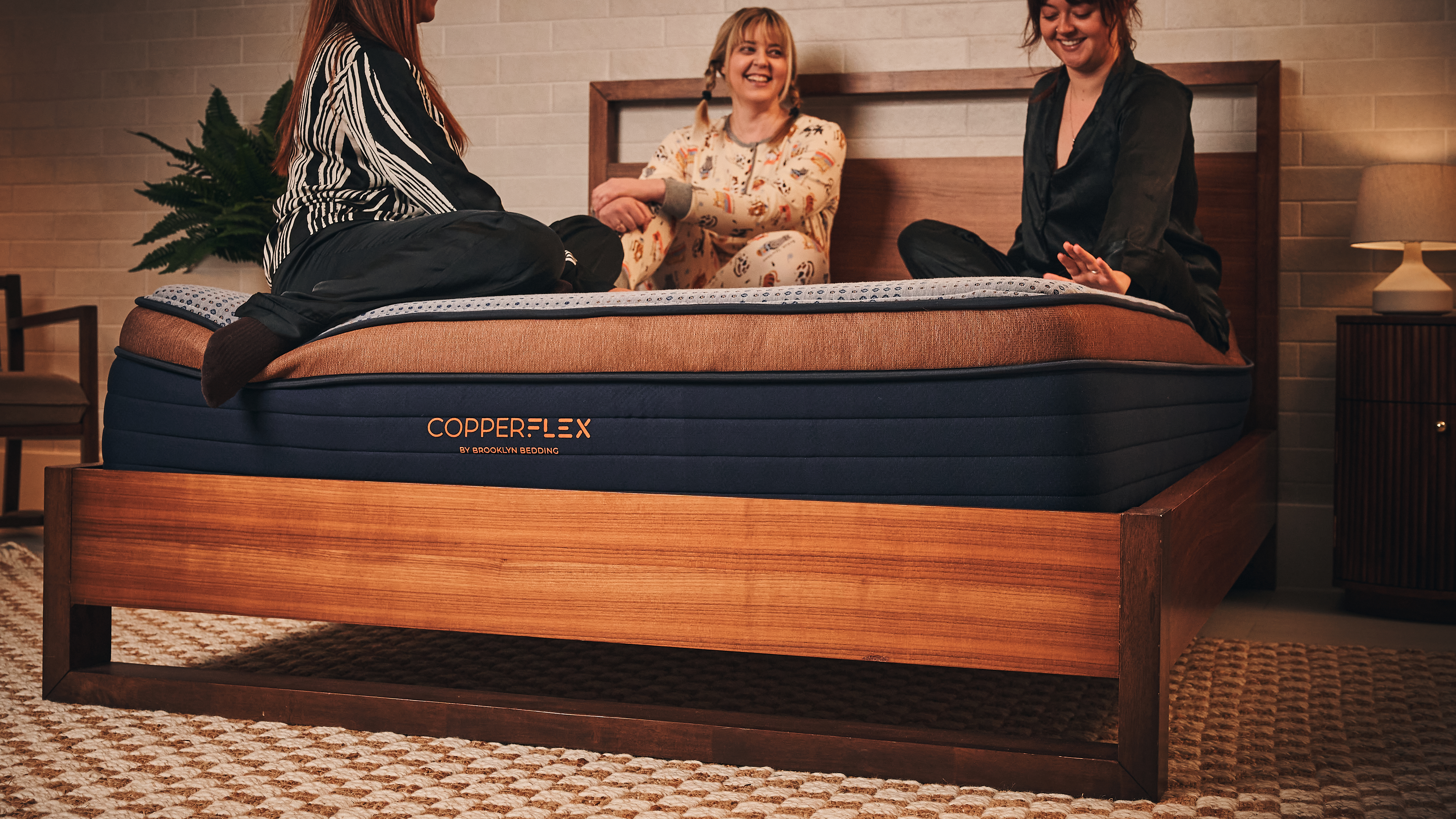 Three women laugh and joke while sitting on a good-quality mattress in a modern, stylish bedroom setting.
