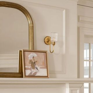 A gold sconce above a white fireplace next to a picture frame and mirror