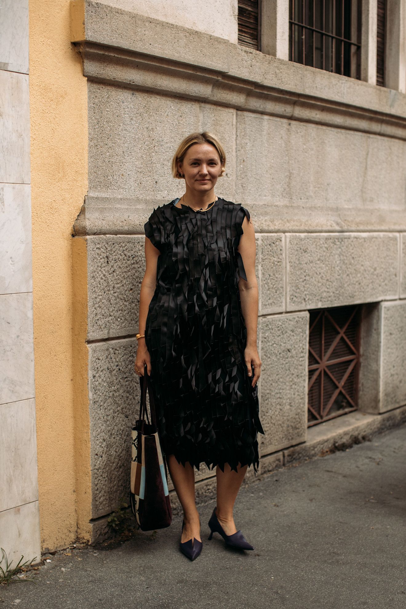 Menswear SS26 showgoer wears a black textured dress with pointed toe black shoes with a multi-coloured tote bag.
