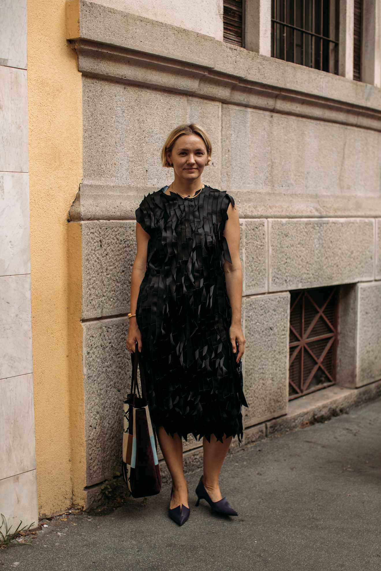 Menswear SS26 showgoer wears a black textured dress with pointed toe black shoes with a multi-coloured tote bag.