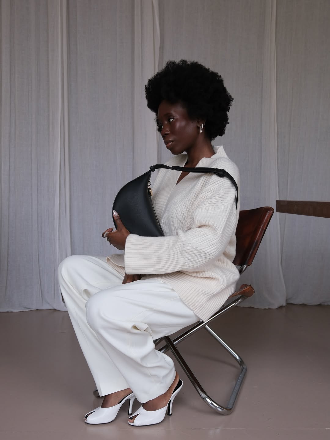 A woman wears white pants with a cream sweater, white peep-toe heels, and a black shoulder bag.