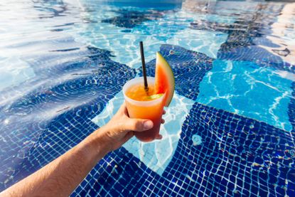 a hand holding an orange cocktail drink in a swimming pool