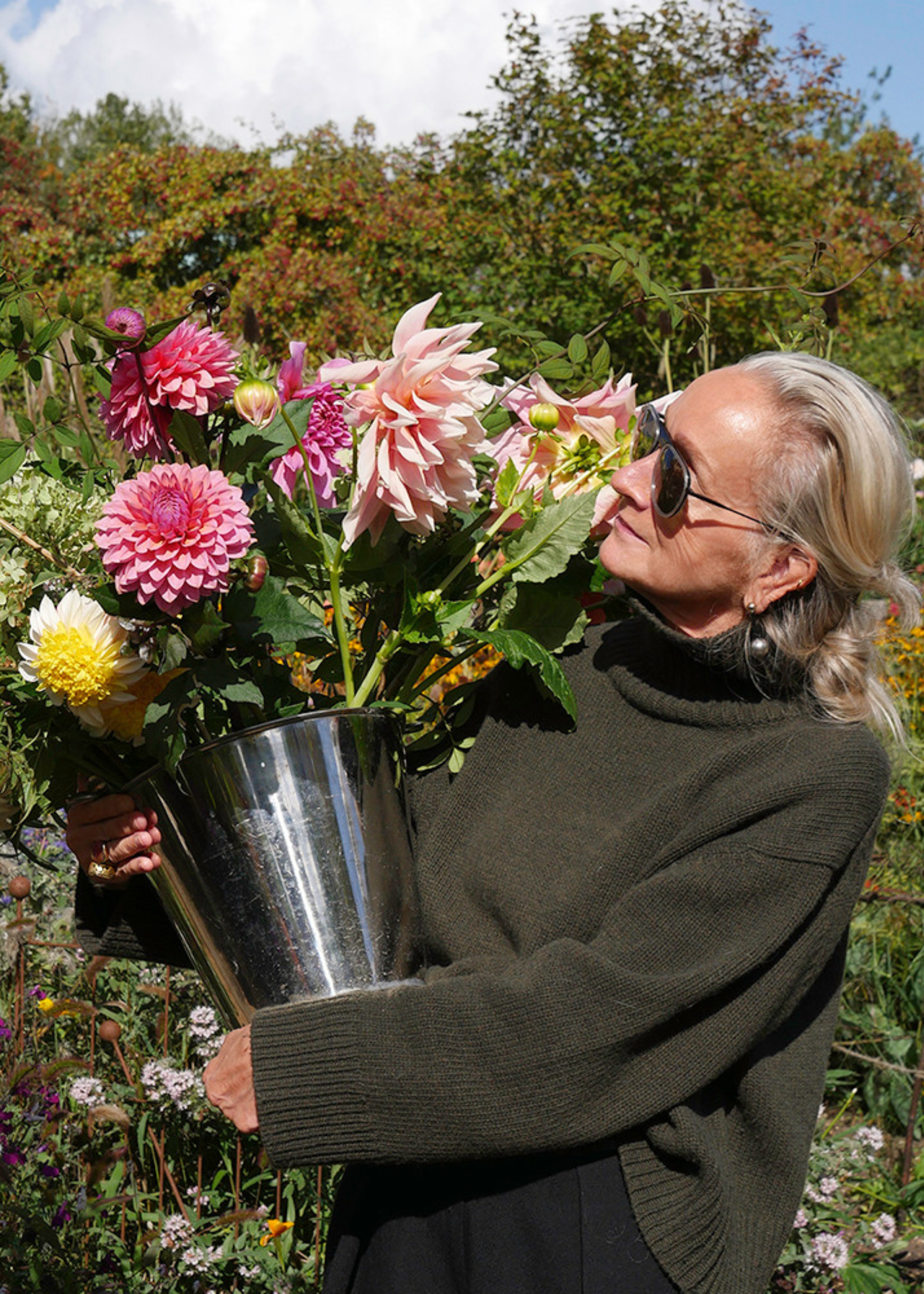 A woman in a turtleneck dark green sweater and aviator sunglasses holding a steel bucket of pink, red, and white and yellow dahlias