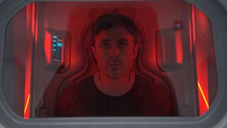 a man with short brown hair looks out of a glass enclosure bathed in red light