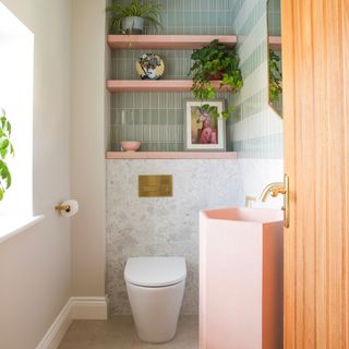 small bathroom with green tiles, pink shelves, marble style tiles on lower wall and floor, brass fixtures and fittings, pink basin