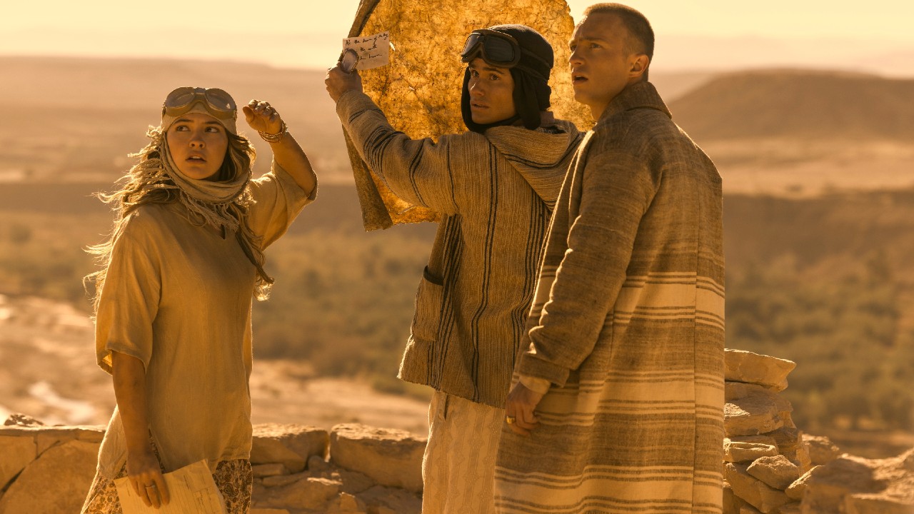 From left to right: Madelyn Cline wearing a head scarf and looking over her shoulder, Chase Stokes holding up a map and looking over his shoulder and Drew Starkey looking over his shoulder.