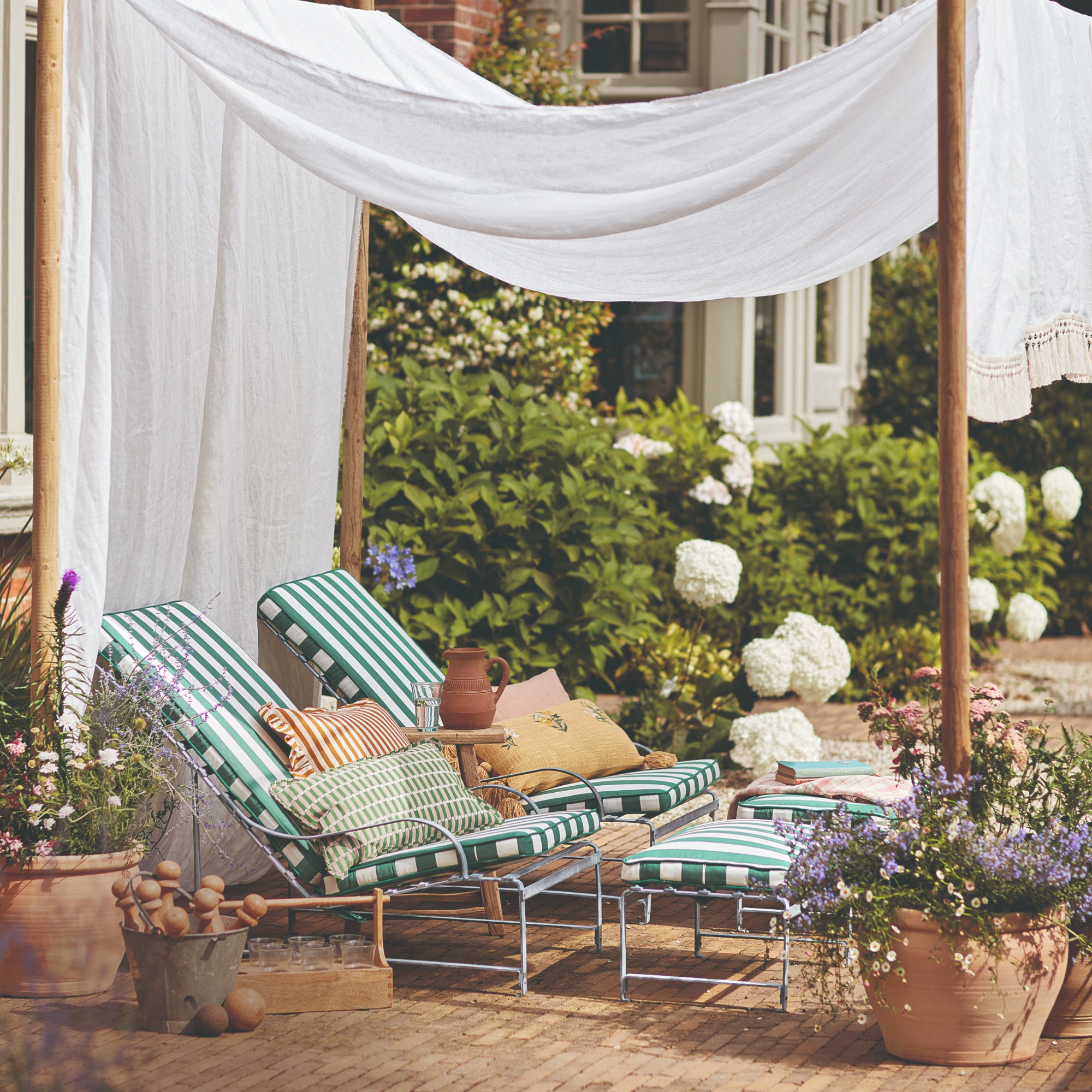 A Mediterranean-style patio with two matching reclined metal garden chairs with striped cushions and matching footstools and a white canopy over them