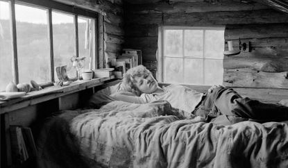 Black and white image showing a woman lying on a bed in a wooden cabin