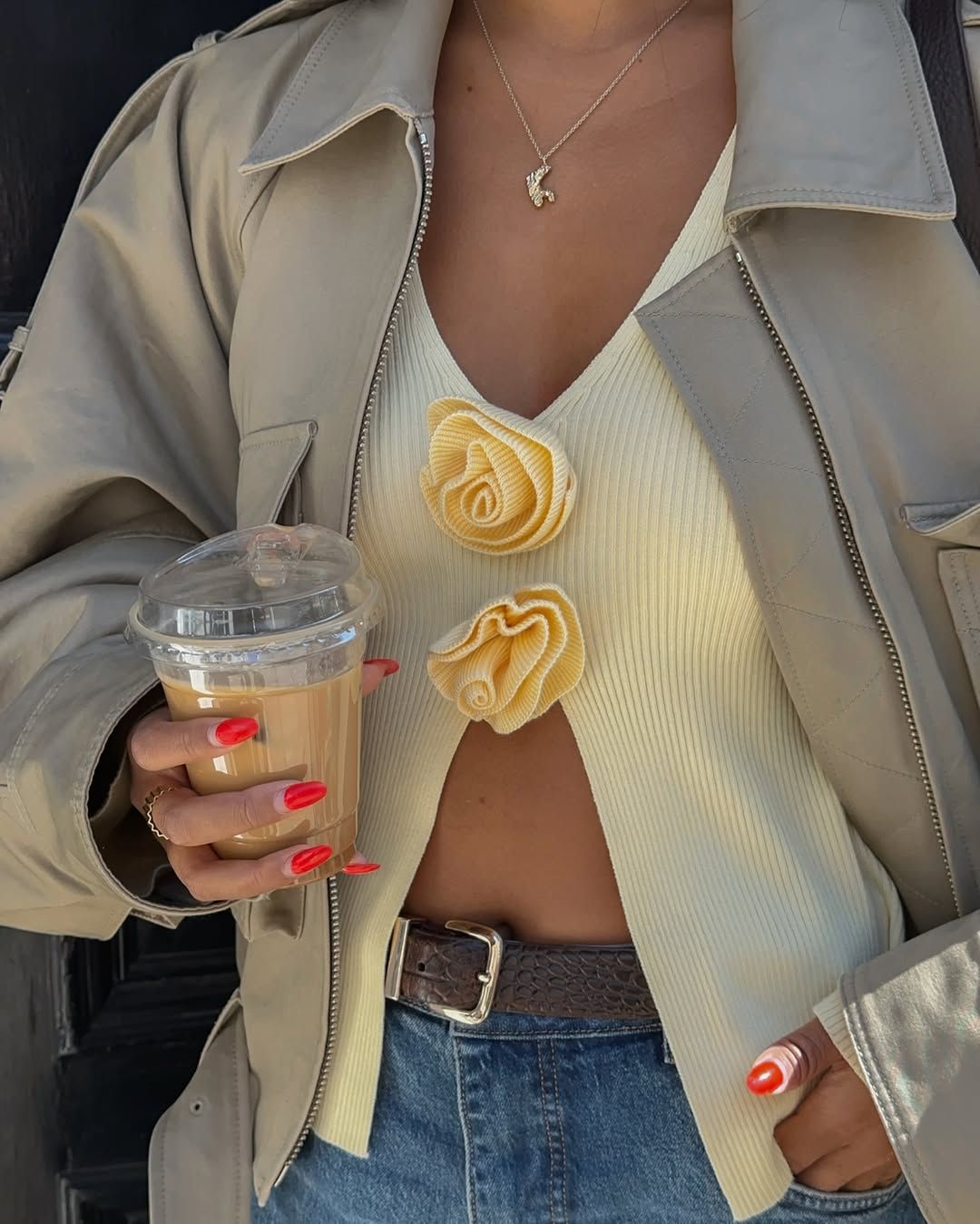 Influencer in a blue jeans, butter yellow flower top, cropped trench holding a coffee with red nails.