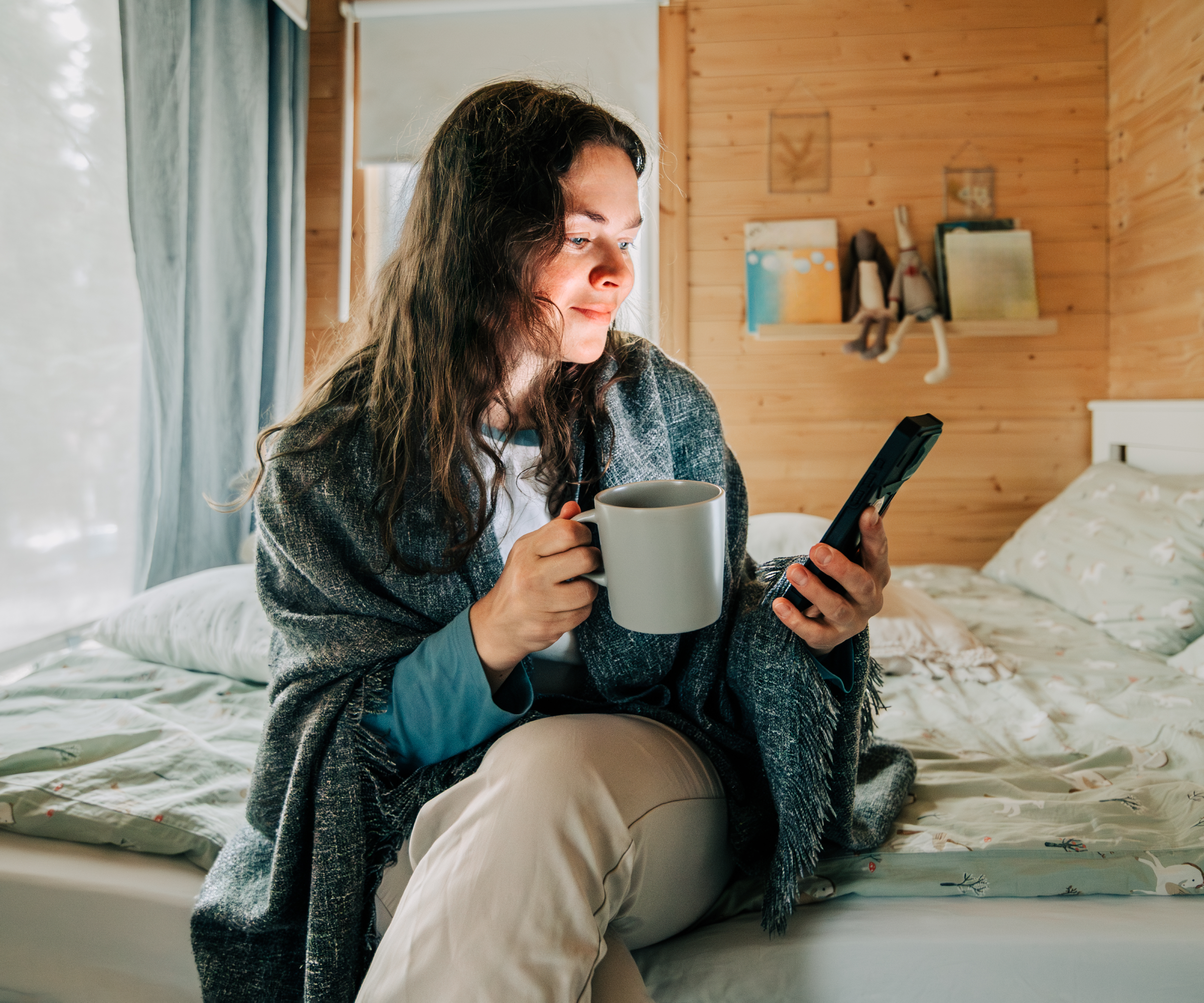 female sat on bed looking at phone with blanket around shoulders and mug in hand