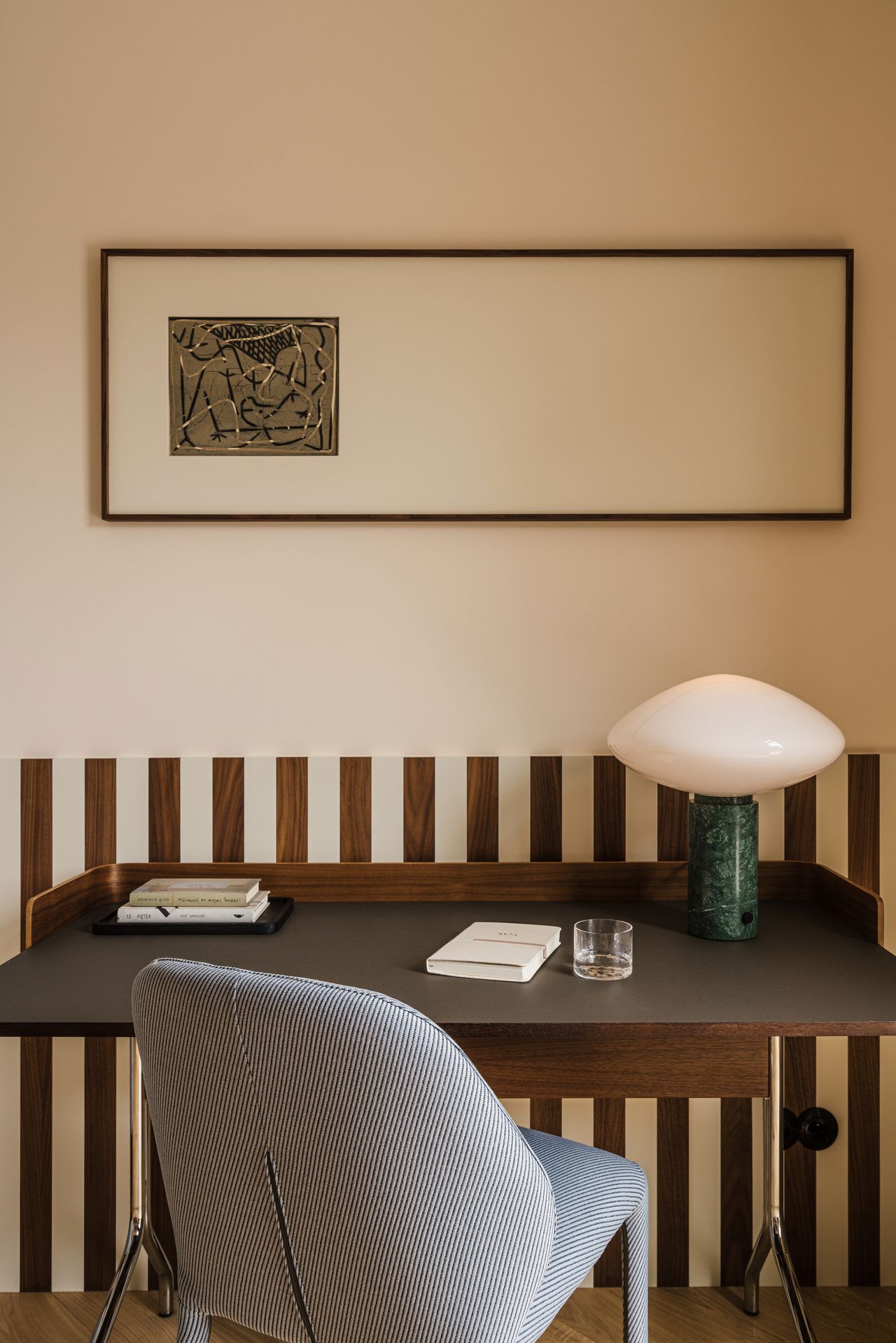Image of a brown desk with a light blue desk chair. The walls are a pinkish-beige with the bottom half being a brown stripe. There is a long, horizontal artwork on the wall and a green and white mushroom lamp on the desk.