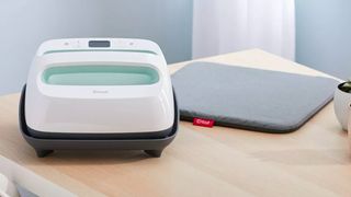 The best Cricut machines; a square craft machine on a black plastic stand, on a wooden table with a grey heat resistant mat