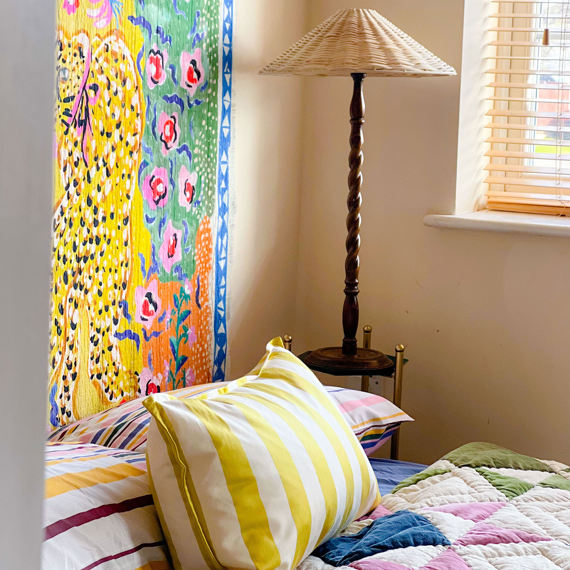 Bedroom corner with tall wooden table lamp and colourful print behind bed