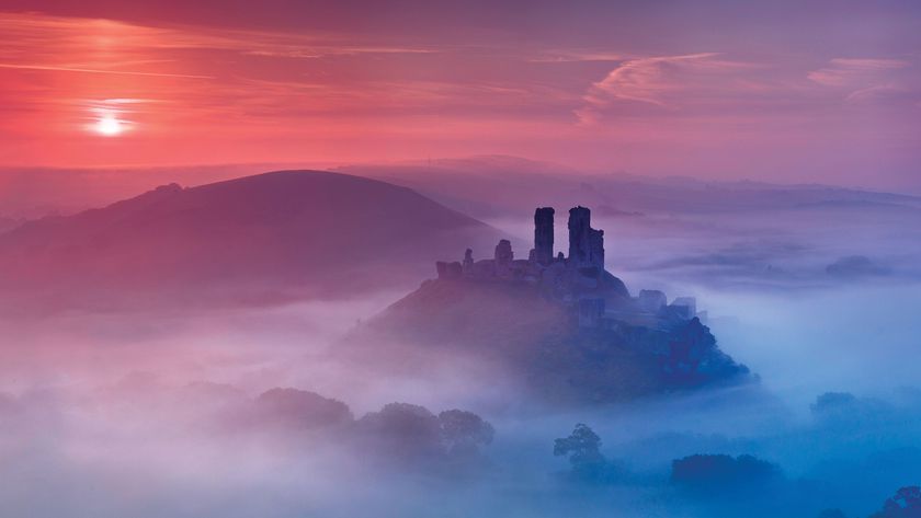 Misty dawn over ruins on a hilltop, surrounded by a foggy landscape. A vibrant sky transitions from deep pink to soft purple, evoking a tranquil mood