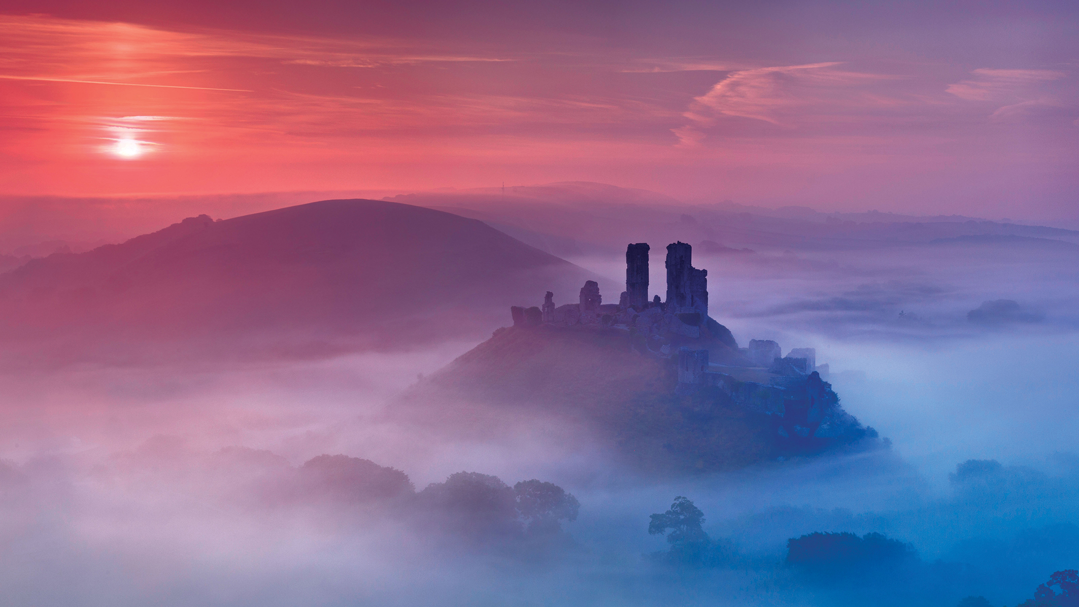Misty dawn over ruins on a hilltop, surrounded by a foggy landscape. A vibrant sky transitions from deep pink to soft purple, evoking a tranquil mood