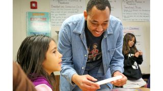 A smiling man in a denim blue shirt helps a young female student with a STEM project in a classroom. 