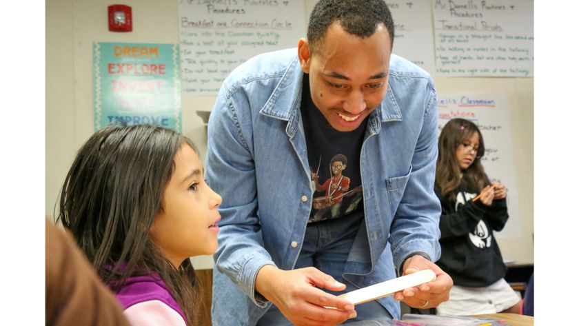 A smiling man in a denim blue shirt helps a young female student with a STEM project in a classroom. 