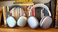 Purple AirPods Max 2 and blue AirPods Max (2024) headphones standing next to each other on a bookshelf in front of a row of books.