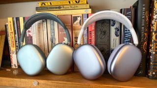 Purple AirPods Max 2 and blue AirPods Max (2024) headphones standing next to each other on a bookshelf in front of a row of books.