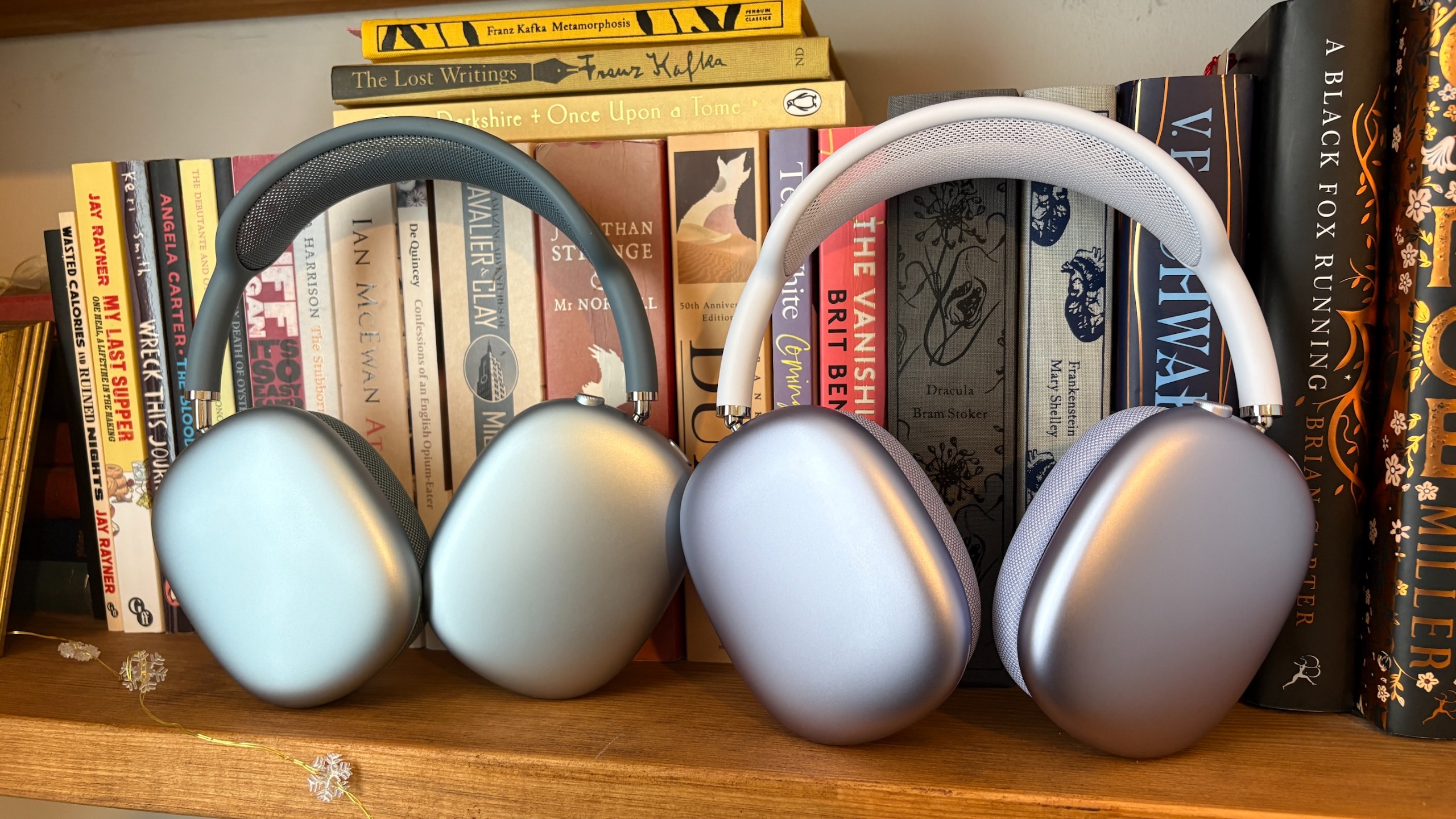 Purple AirPods Max 2 and blue AirPods Max (2024) headphones standing next to each other on a bookshelf in front of a row of books.